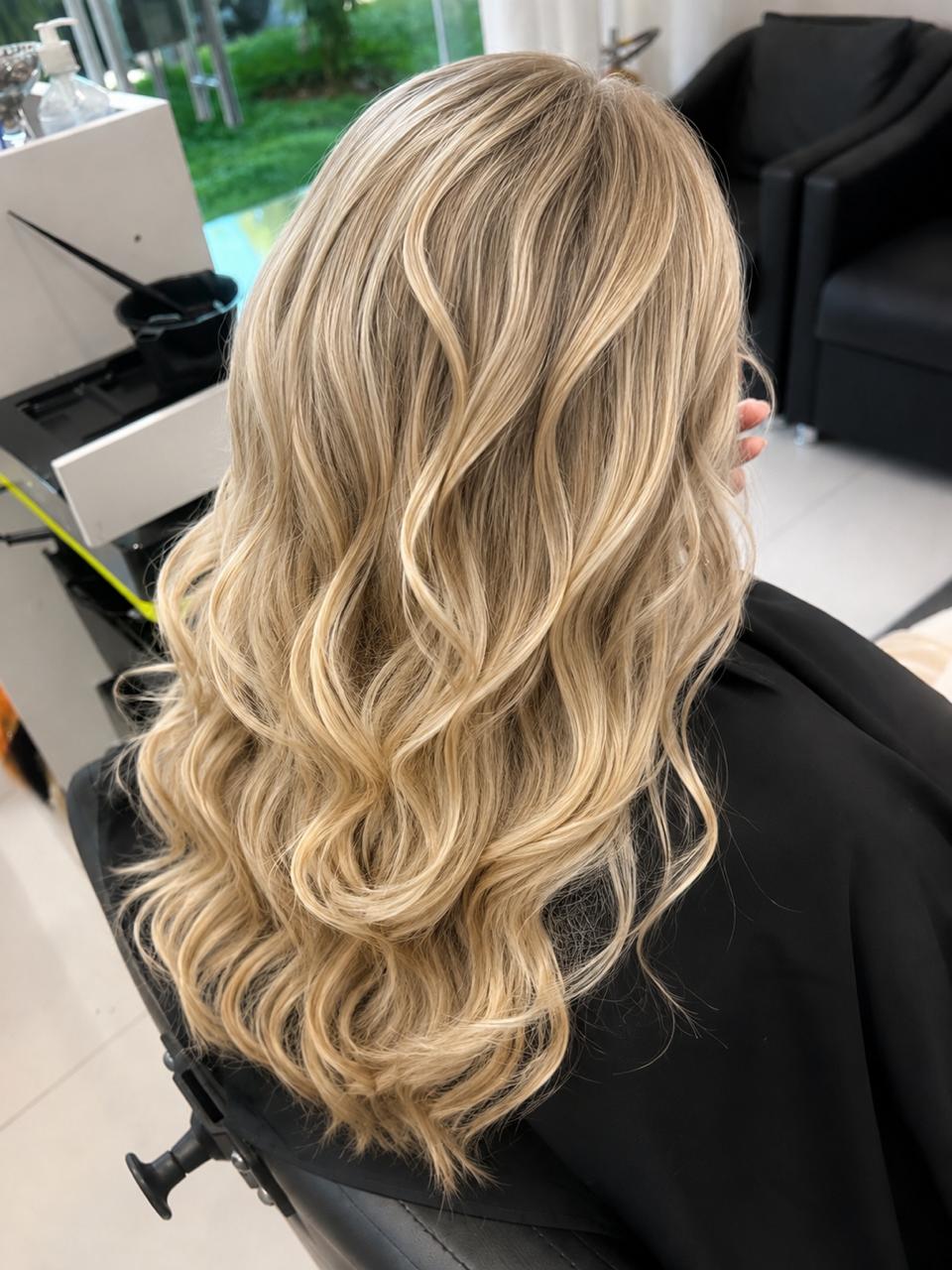 Resultado de mechas em loiro multidimensional com tons mel e acinzentado, cabelo longo com ondas soltas, visto de costas no salão.
